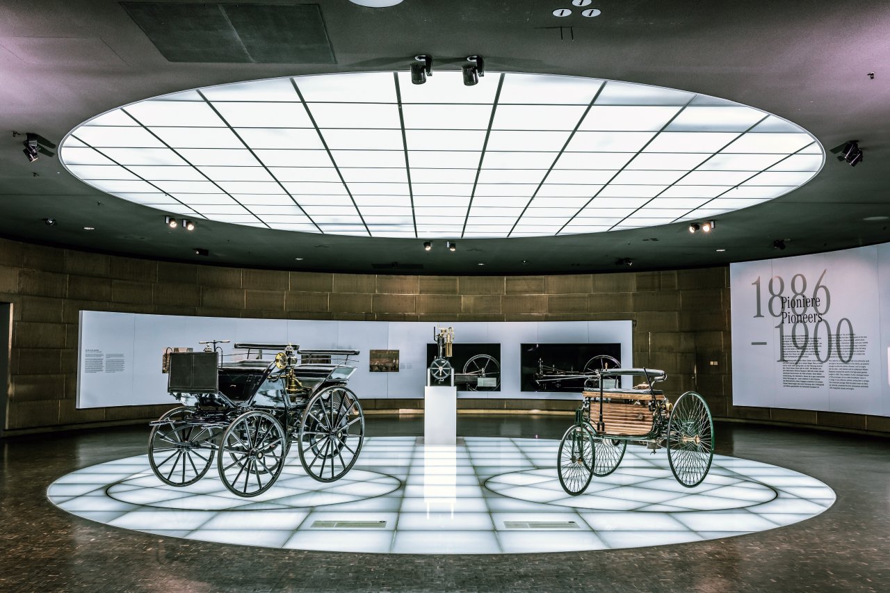 Benz Patent-Motorwagen (desno) in Daimler Motorkutsche (levo) na razstavnem prostoru, ki zajema obdobje od leta 1886 do 1900. V središču je Daimlerjeva »stoječa ura« – prvi majhen, hitrovrteči se motor z notranjim zgorevanjem.